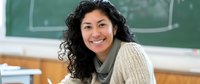 Young Hispanic female teacher with curly dark hair and warm smile sitting in classroom with chalkboard background, wearing casual sweater and scarf.