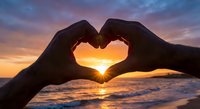 Hands forming heart shape silhouette against golden sunset over ocean waves. Romantic gesture symbolizing love, connection and unity at beach during twilight.