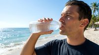 Man drinking water from bottle on tropical beach with ocean and palm trees in background, staying hydrated during summer vacation or outdoor activities.