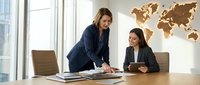 Two professional businesswomen collaborating in modern office with world map, discussing strategy and documents at conference table near windows.