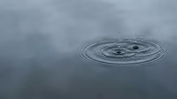 Water droplet creating concentric ripples on calm gray surface with cloudy sky reflection, symbolizing tranquility, meditation and peaceful moments in nature.