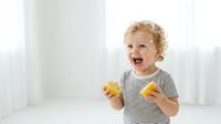 Toddler with curly blonde hair, Caucasian ethnicity, wearing striped shirt, laughing while holding orange slices in bright, clean studio setting.