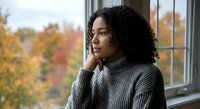 Thoughtful African American woman in gray sweater gazing out window during autumn season, reflecting on life decisions and future possibilities.