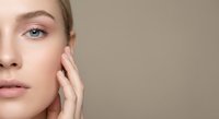Young Caucasian woman with natural makeup touching face showing healthy glowing skin for skincare and beauty treatments on neutral background.