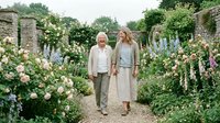 Senior couple strolling through blooming garden with roses and flowering plants, enjoying outdoor leisure time together in romantic botanical setting.