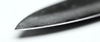 Sharp kitchen knife blade tip in extreme close-up macro shot, showing steel texture and cutting edge detail on white background.