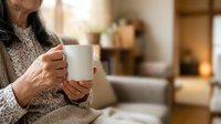 Senior woman with wrinkled hands holding white mug in cozy living room, wearing warm sweater. Use for lifestyle, wellness, comfort, home relaxation, and beverage marketing campaigns.