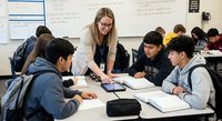 Young blonde female teacher with glasses helping diverse high school students with assignments in modern classroom setting during collaborative learning session.