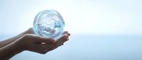 Close-up of hand holding transparent globe filled with water against blue sky background, symbolizing environmental conservation and sustainability.