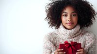 Young African American woman with natural curly hair wearing cozy turtleneck sweater holds red gift box with bow against white background, expressing joy and gratitude.