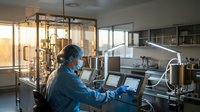 Female scientist in protective mask working at computer in modern pharmaceutical laboratory with stainless steel equipment and glass partitions during research.