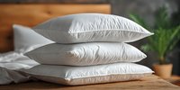 Stack of three white fluffy pillows on wooden surface with green plant in background for bedroom and home decor concepts.