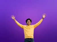 Man with dark curly hair wearing bright yellow hoodie celebrating with arms raised against vibrant purple background, expressing joy and success for lifestyle and motivation concepts.