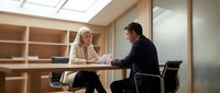 Mature woman with blonde hair and middle-aged man in business attire having professional discussion at table in modern office setting with wooden shelving.