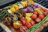 Grilled colorful vegetables on wooden cutting board including asparagus, bell peppers, onions, and cherry tomatoes with charred grill marks and seasoning.