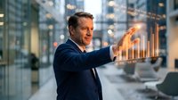Middle-aged Caucasian businessman in navy suit interacting with holographic financial charts and data visualization in modern corporate office environment.