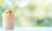 Creamy caramel frappe topped with whipped cream and drizzle in clear plastic cup against soft green bokeh background, refreshing summer drink.
