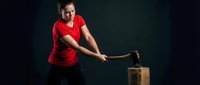 Young athletic woman in red shirt swinging sledgehammer at wooden box during crossfit training workout on dark background for fitness motivation.
