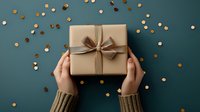 Hands holding elegant kraft paper gift box with brown bow against dark green background with golden confetti. Festive holiday present concept.
