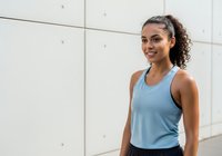 Young woman with curly dark hair and olive complexion wearing light blue athletic tank top, smiling at camera in modern gym or fitness studio with white paneled walls. Fitness and wellness.