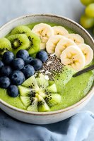 Fresh green smoothie bowl topped with sliced kiwi, banana, blueberries and chia seeds. Healthy breakfast with tropical fruits served in ceramic bowl.
