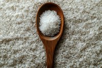 Wooden spoon filled with coarse sea salt crystals on textured white background, top view composition for culinary and wellness concepts.