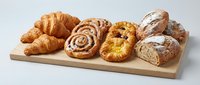Assorted fresh baked pastries and bread on wooden board including croissants, cinnamon rolls, and artisan loaves for bakery breakfast menu display.