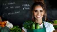 Young female chef holding fresh broccoli and cucumber in modern kitchen with chalkboard menu background for healthy cooking and restaurant concepts.
