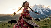 Young Tibetan woman in traditional colorful dress dancing joyfully in mountain meadow at sunset with yaks grazing in background landscape.