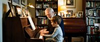 Senior man with white hair teaching young boy piano in home library with bookshelves, warm lighting, intergenerational music lesson and family bonding moment.