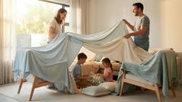 Happy family building blanket fort at home with children playing underneath while parents hold sheet creating cozy hideout in bright living room.