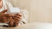 Mature adult with dark skin tone holding blank white business card on wooden surface, wearing casual beige clothing, suitable for corporate branding and professional networking materials.