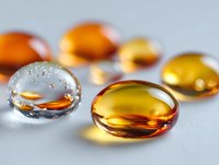 Transparent gel capsules with orange and yellow liquid medicine on light background, macro photography showing water drops and reflective surfaces.
