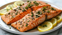 Grilled salmon fillets seasoned with herbs and black pepper, served on white plate with fresh lemon slices and garnished with chopped parsley.