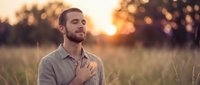 Man with hand on chest standing in golden wheat field at sunset, expressing gratitude, peace, or mindfulness in rural agricultural setting.