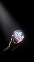 Business hand holding glowing target icon with arrow in center against dark background representing goal achievement and strategic planning success.