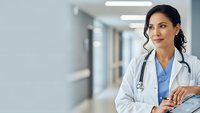 Confident female doctor with stethoscope standing in modern hospital corridor wearing white coat and blue scrubs for healthcare marketing.