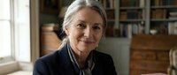 Professional mature Caucasian businesswoman with gray hair smiling confidently in modern office environment with bookshelves and warm natural lighting.