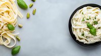 Fresh fettuccine pasta with creamy sauce, scattered pistachios and basil leaves on light marble background, top view with copy space for menu design.