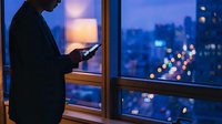 Businessman using smartphone by office window at night with city lights bokeh background. Professional working late hours in modern corporate building.