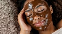 Young African American woman enjoying chocolate face mask spa treatment with closed eyes and serene smile, lying on towel during skincare routine.