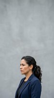 Professional woman with dark hair in ponytail, wearing navy blazer, photographed in profile against neutral gray concrete wall background for corporate and business use.