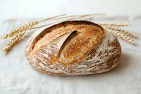 Fresh artisanal sourdough bread loaf with crispy crust and flour dusting on white linen cloth, decorated with golden wheat stalks, rustic bakery setting.