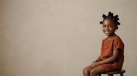 Young African girl with black braided hair styled with decorative clips, wearing a rust-colored shirt, smiling at camera while seated against neutral background for portrait and lifestyle use.