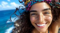Young mixed race woman with curly hair and colorful headband smiling brightly at beach, natural freckles, close up portrait against blue ocean and sky background.
