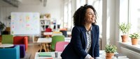 Young woman with curly dark hair and medium skin tone, wearing navy blazer, sitting in modern office space with colorful furniture and plants, looking thoughtful while working on business project.