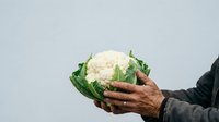 Mature hands holding fresh white cauliflower with green leaves against neutral background, suitable for organic food, agriculture, healthy eating, and farm produce marketing materials.
