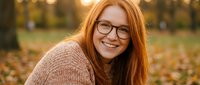 Young redhead woman with glasses smiling warmly in autumn park during golden hour. Natural portrait for lifestyle, wellness, happiness concepts.