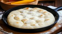 Raw focaccia dough with dimples in black cast iron pan, dusted with flour, ready for baking, traditional Italian bread preparation on wooden surface.