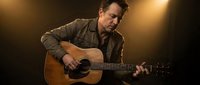 Middle-aged Caucasian male musician playing acoustic guitar on stage with dramatic warm lighting, professional concert performance setting.